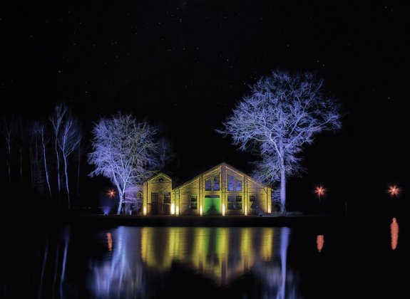 Die alte Werkshalle der ehemailigen Dynamitfabrik auf dem Campus Nobel in Saarwellingen. Die Backsteinhalle ist mit gelben und blauen Lichtern angestrahlt. Es ist Nacht. Die Halle spiegelt sich im davor gelegenen See.